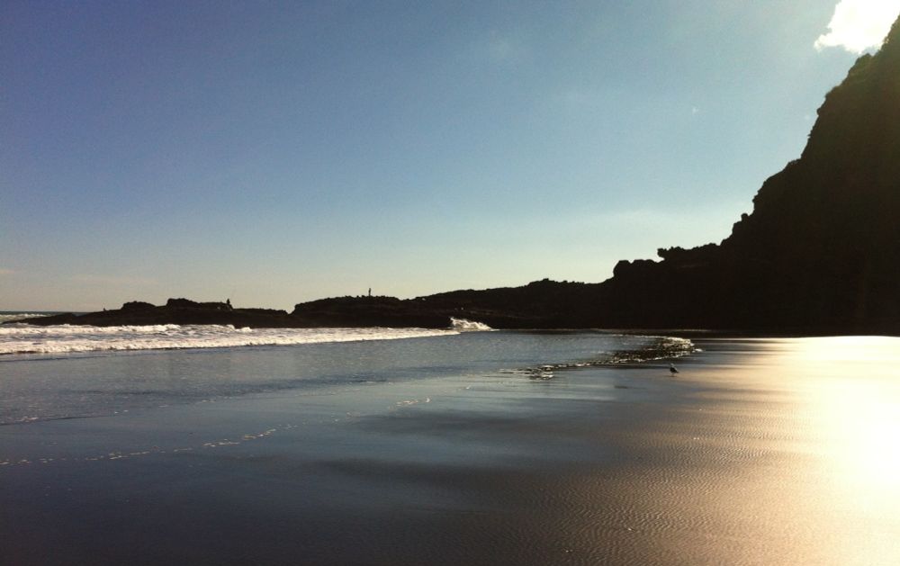 From my old iPhone 4 - July 2014.  One of the wild West Coast beaches is aglow with light. The sun is diffused thru a cloud in the upper right-hand portion of the photo. The sand is near flawless, no footsteps - pristine and smooth. It reflects the glow from above. Horizontally dissecting the middle of the photo is a rocky outcrop that juts out into the Tasman. Fisherman are on the rocks working their craft. A lone seagull is on the sand watching the waves.  