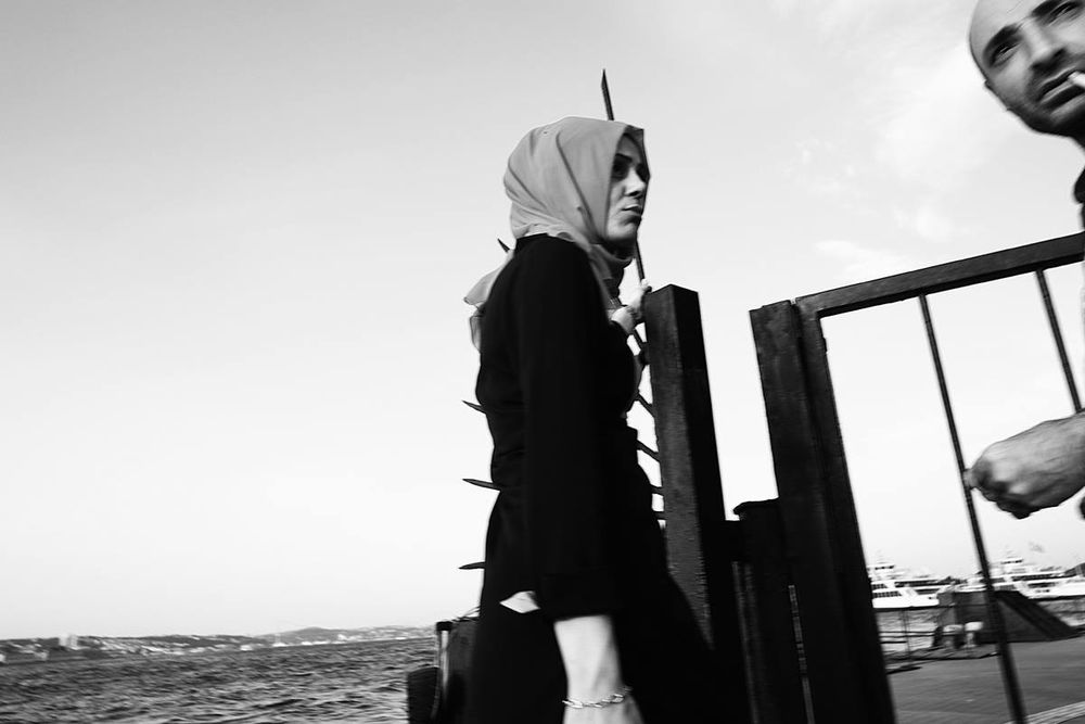 Cette photographie de rue en noir et blanc, probablement prise à Istanbul, capture un moment fugitif empreint de tension visuelle et émotionnelle. Voici une description détaillée :

Sujet principal :
	•	Une femme au premier plan vêtue d’un manteau sombre et portant un hijab clair (probablement rose ou beige en réalité, mais ici en nuances de gris), traverse la scène de profil. Son visage est sérieux, presque fermé, et elle semble absorbée dans ses pensées ou dans une forme de mélancolie.
	•	Un homme en arrière-plan à droite, partiellement cadré, regarde vers l’objectif avec un regard perçant. Il tient une cigarette à la bouche, ce qui ajoute une certaine rudesse ou nonchalance à son apparence. Il est flou, ce qui accentue l’effet de mouvement ou de saisie spontanée.

Composition :
	•	L’image est coupée de façon asymétrique, créant une tension dynamique entre les deux personnages.
	•	Des barres de métal (clôture ou grille) et des pointes métalliques acérées en arrière-plan viennent accentuer l’impression d’isolement ou d’oppression.
	•	À gauche, on aperçoit la mer de Marmara ou le Bosphore, avec un horizon lointain et des bâtiments indistincts, qui évoquent Istanbul.
	•	Des bateaux amarrés apparaissent à droite, suggérant un port ou une jetée, élément typique du paysage stambouliote.

Atmosphère :
	•	Le noir et blanc renforce l’effet dramatique et le contraste entre les zones claires (ciel, mer) et les zones sombres (vêtements, structures métalliques).
	•	L’image évoque un instant volé, typique de la photographie de rue, où les sujets ne posent pas mais sont saisis dans leur quotidien.
	•	Il y a une dualité forte entre la femme qui semble introvertie, presque absente, et l’homme dont le regard est frontal, presque intrusif.

Lecture possible :

Cette scène peut symboliser la cohabitation des tensions sociales, culturelles ou de genre dans l’espace public à Istanbul.