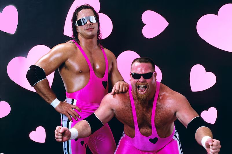 The Hart Foundation featuring Bret "The Hitman" Hart and Jim "The Anvil" Neidhart in all pink wrestling attire posed in front of a wall with large pink hearts on a black background.