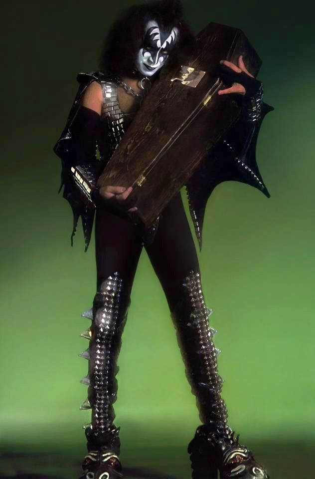 This photo is of Gene Simmons in 1977 in full costume and signature makeup with a green background. He's cradling a small 3-foot wooden coffin in his arms while he shoots a sinister glare at the camera.
