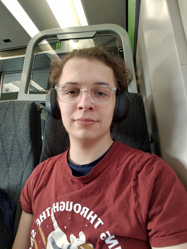 White individual with clear glasses wearing headphones looking directly at the camera while sat in a train.