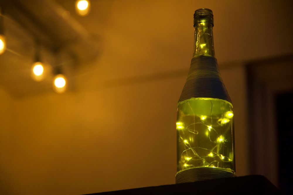 photo of a green glass bottle wrapped with twine on the base and neck filled with fairy lights. The bottle sits to the right of frame and the camera is looking up at it with a white building in the background and some warm lights to the top left illuminating the background.