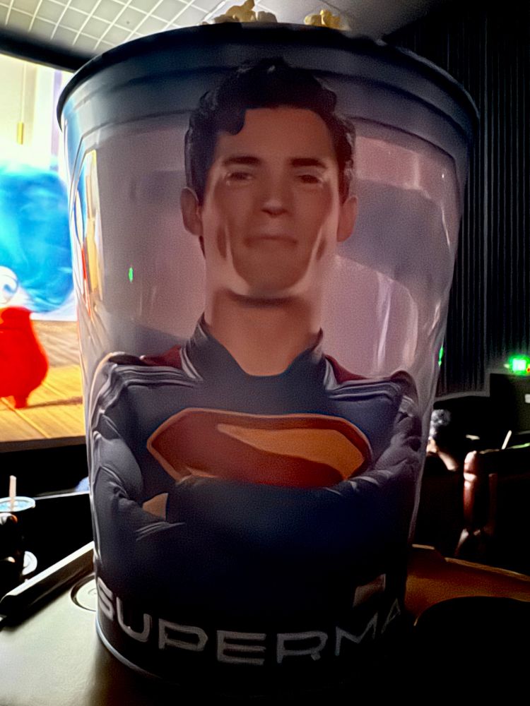 A bucket of popcorn in a movie theater. The exterior of the bucket has a photo on it of Clark Kent wearing a Superman costume.