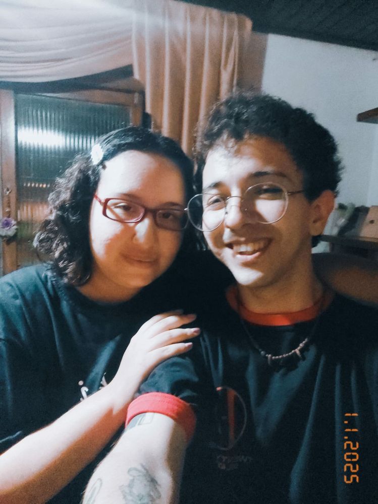 mulher e homem ambos de cabelos escuros e cacheados, vestindo camisetas pretas. a mulher usa óculos quadrados vermelhos e o homem óculos redondos e metálicos.