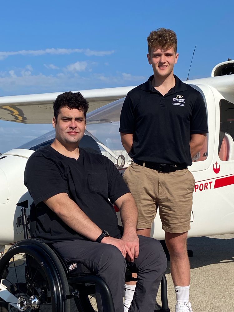 Student pilot Tim Riney, Jr. alongside his flight instructor Billy O’Connor during Able Flight’s 2024 training program at Purdue University. A red and while two seat training plane is behind them. During that program Tim earned his pilot’s license. 

Tim uses a wheelchair since becoming paralyzed during a training accident while serving in the Army. 