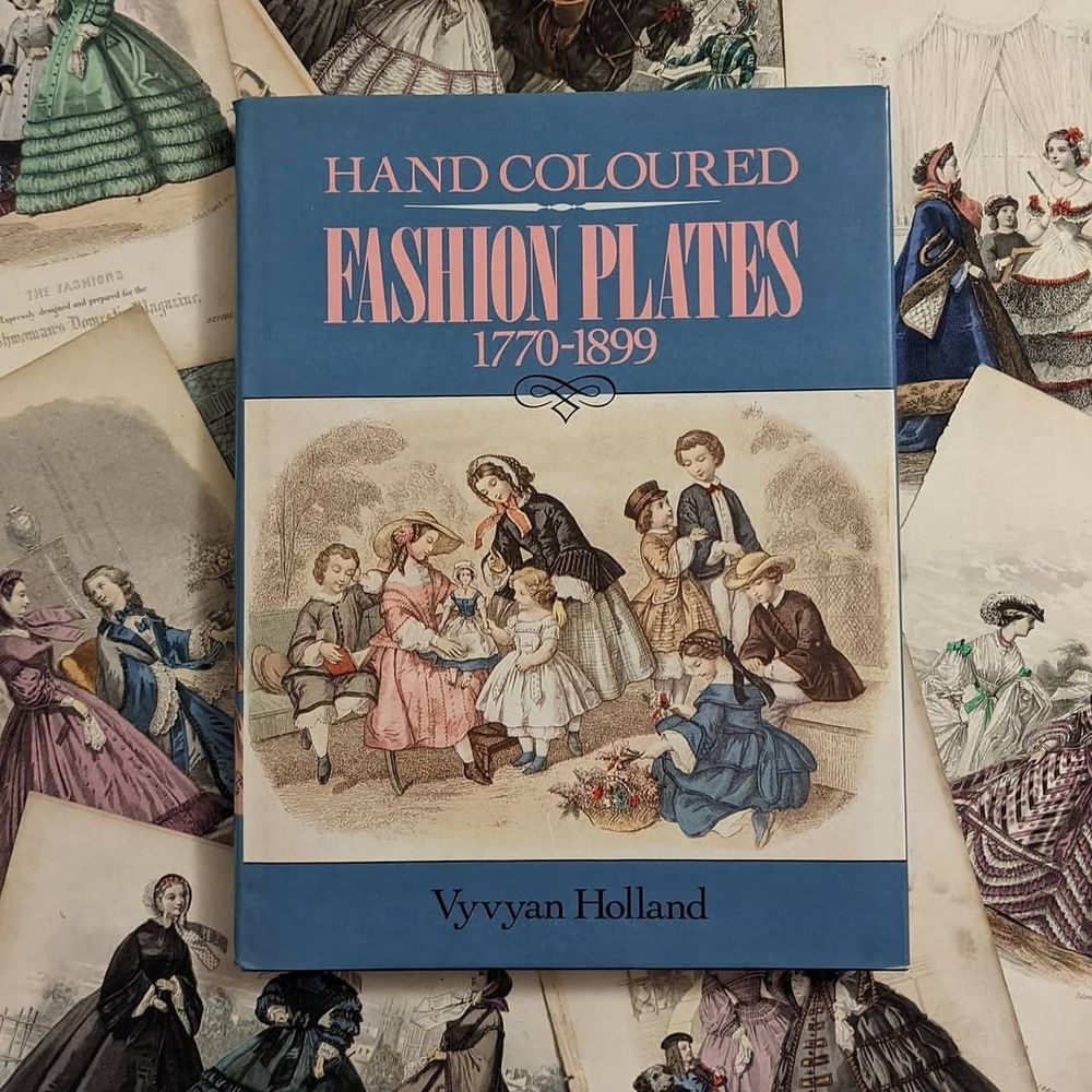 A book titled Hand-Coloured Fashion Plates 1770-1899 by Vyvyan Holland, lying on top of a selection of 1860s fashion prints which are only partially seen around the outside of the book.

Photo taken by Daniel Milford-Cottam, 2021
