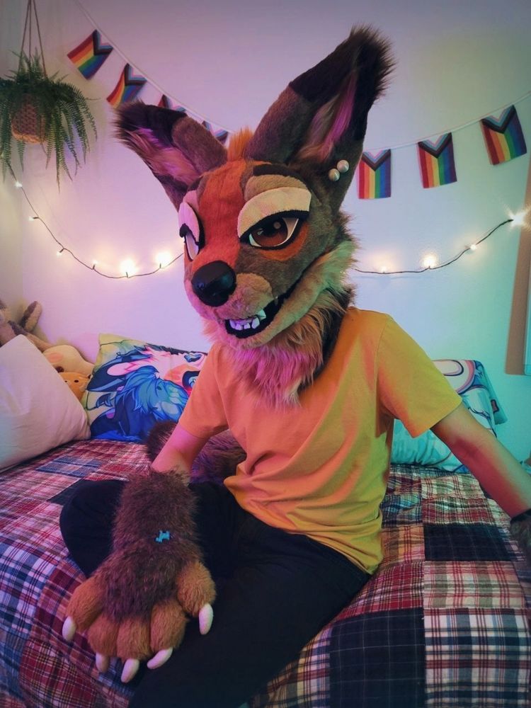 A photo of a coyote partial fursuiter, sitting on a bed and smiling at the camera