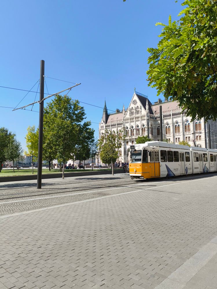 Clean and well put together public area with a lot of trees and a street train passing by. People enjoying their day on benches and walking around 