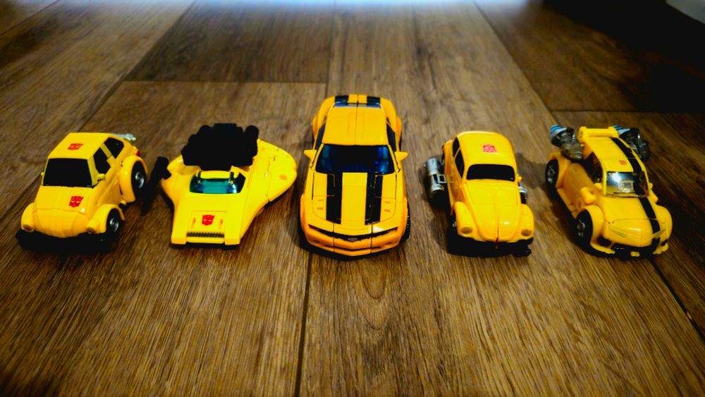 A collection of Transformers Bumblebee toys in vehicle mode. Left to right: Studio Series 86 Bumblebee (a yellow penny racer type car), Origins Bumblebee (a yellow flying saucer), Camaro Concept Bumblebee, Retro G1 Bumblebee (a yellow VW Beetle), and Animated Bumblebee (a yellow Beetle-style car with rocket boosters).