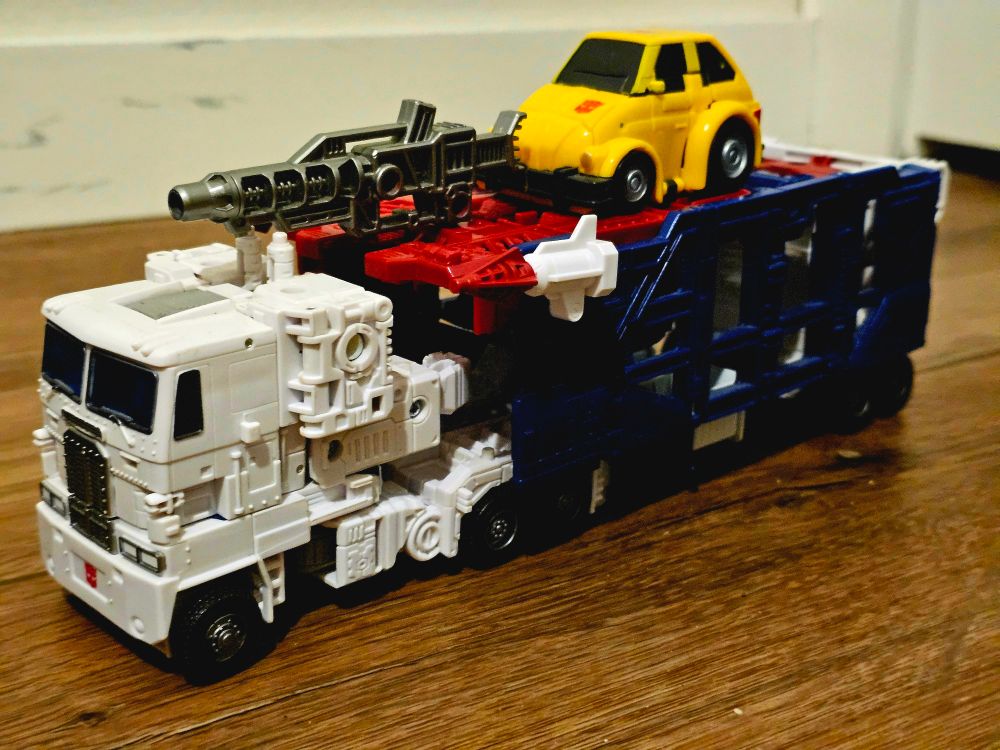 War for Cybertron: Kingdom Ultra Magnus, a Transformers toy that is a white truck with a blue trailer, with missiles and guns attached to his trailer, and Bumblebee, a small yellow robot that transforms into a Volkswagen Beetle, riding on the trailer.