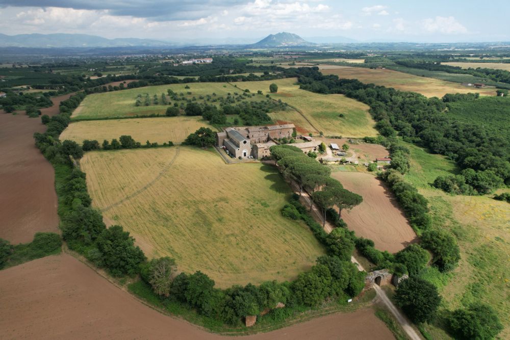 An aerial image of the ancient town of Falerii Novi with wall circuit visible under a tree line, ancient gate in foreground, and a road leading into the site to a church and farmstead. Surrounded by agricultural fields and a mountain in the distance. 
