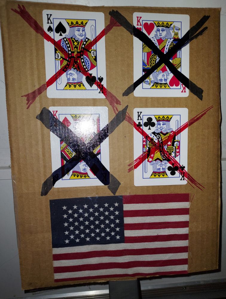 The four kings from a deck of cards are mounted on a piece of cardboard. The kings from black suits are crossed out in red marker, and the kings from red suits are crossed out in black. They appear above a miniature American flag.