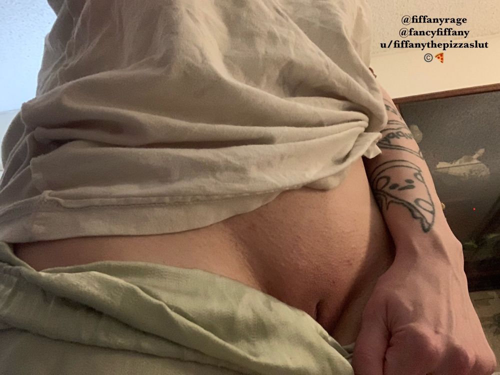 The picture shows the mid section of Fiffany’s body. They wear a large white shirt and green satin pajama pants. They are pulling down their pants with their left hand. You can see that between their legs the skin had been freshly waxed and is smooth
