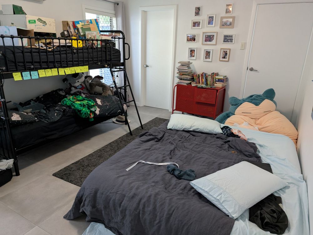 Airbed next to a black bunkbed. The top of the bunkbed is covered in boxes and assorted belongings of a 10 year old boy. The rear wall has a red chest of drawers covered in piles of books, mainly graphic novels. A giant cartoon character beanbag is next to the chest of drawers.