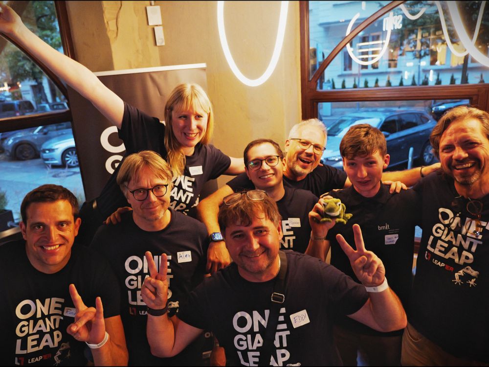Eight people in matching “ONE GIANT LEAP” shirts smile together at a team event. One holds a green frog plush.