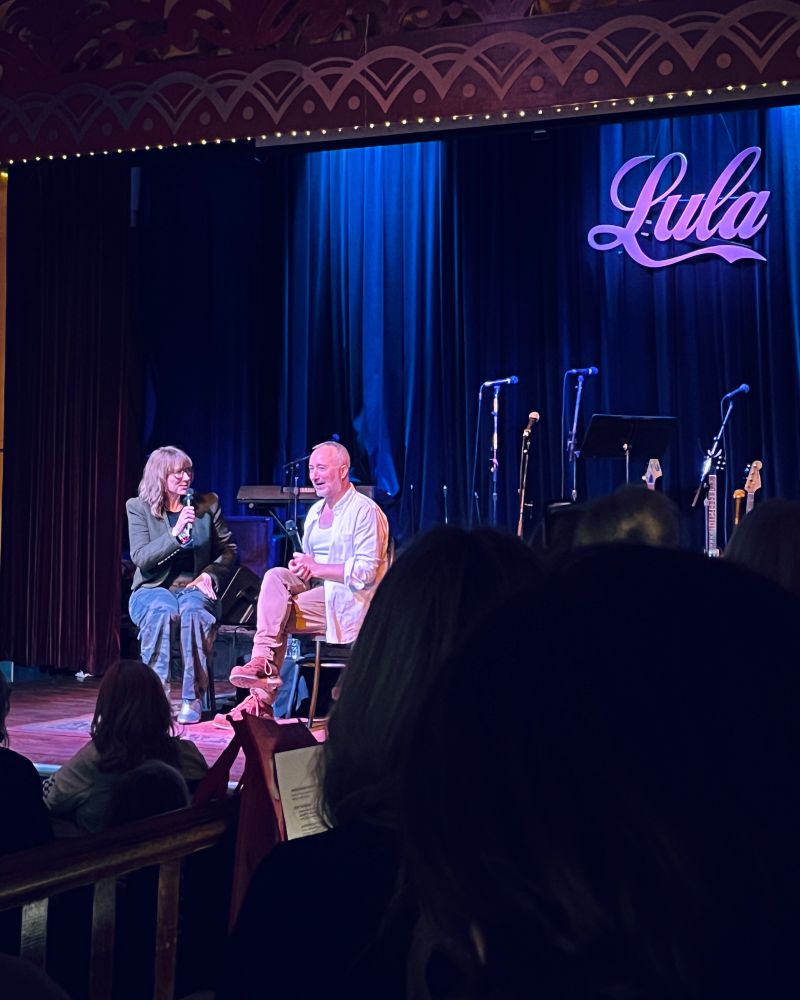 Two people are sitting on a stage in a darkened theatre. 