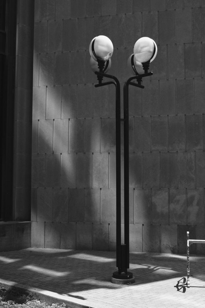 A black and white image of a lamp post with four light globes with angular shadows in the background. 