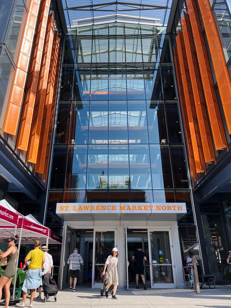 The front doors of the north St Lawrence Market building. Shoppers are browsing stalls and walking about in the foreground. 