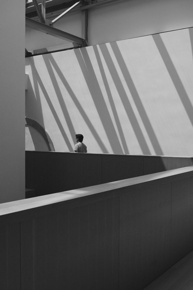 A black and white image of a person walking through striped shadows. There is a railing angled across the foreground. 
