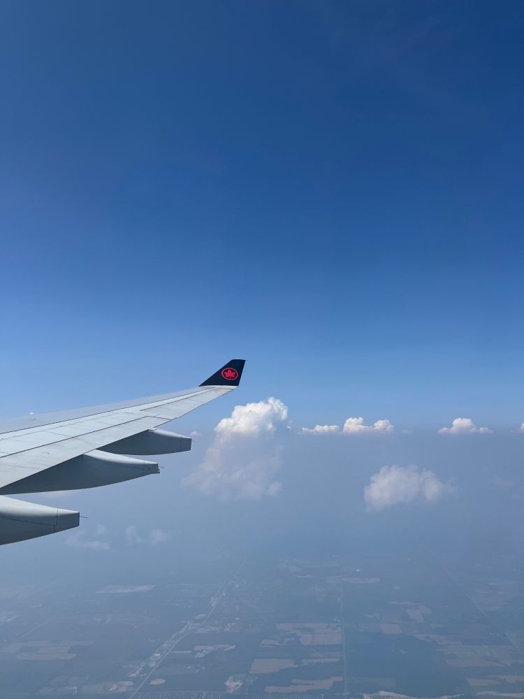 Looking out of a plane window across the wing, there are puffy white clouds in the distance. There is a clear line bisecting the clouds showing hazy smoke on the lower half. 
