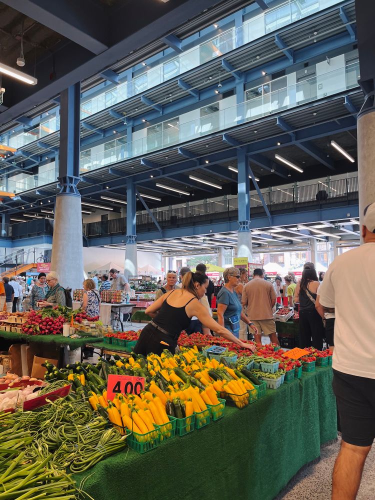 Interior shot of the St Lawrence Market farmers market. A table full of vegetables showing green onions, garlic scapes and zucchini is in the foreground. 