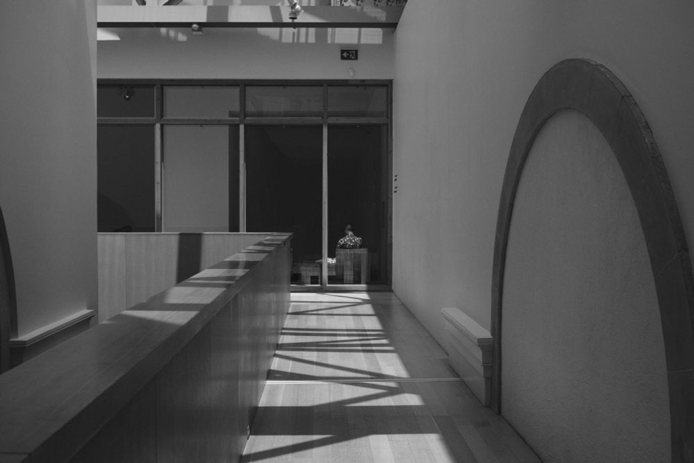 A black and white image along a hallway with striped shadows, a circular shape on the wall and a person visible at the end of the hallway through a window, sitting with their back to the camera. 