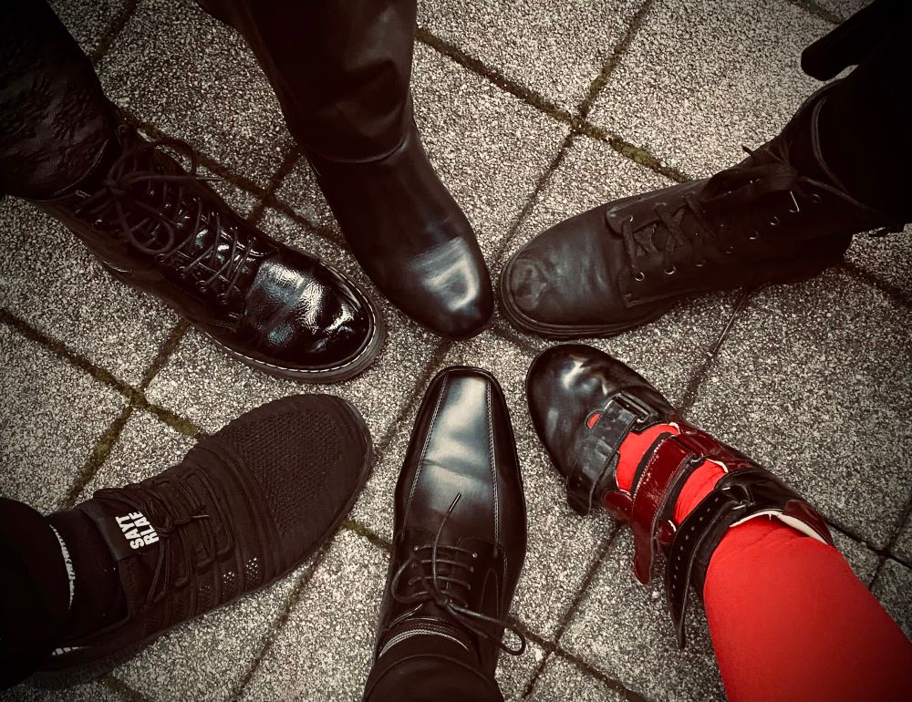 6 Füße in unterschiedlichen Schuhen/Stiefeln, die sich sternförmig an den Zehen treffen…