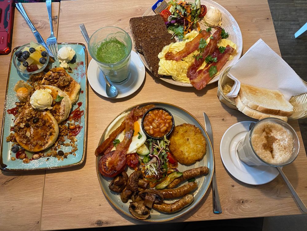 Frühstück superschön angerichtet auf einem Holztisch auf 3 Tellern. Pancakes, Rührei mit Bakon und Vollkornbrot, English Breakfast mit Würstchen, Rösti, Bonen und Spiegelei. Dazu Toast. Dazu zwei Heißgetränke. 