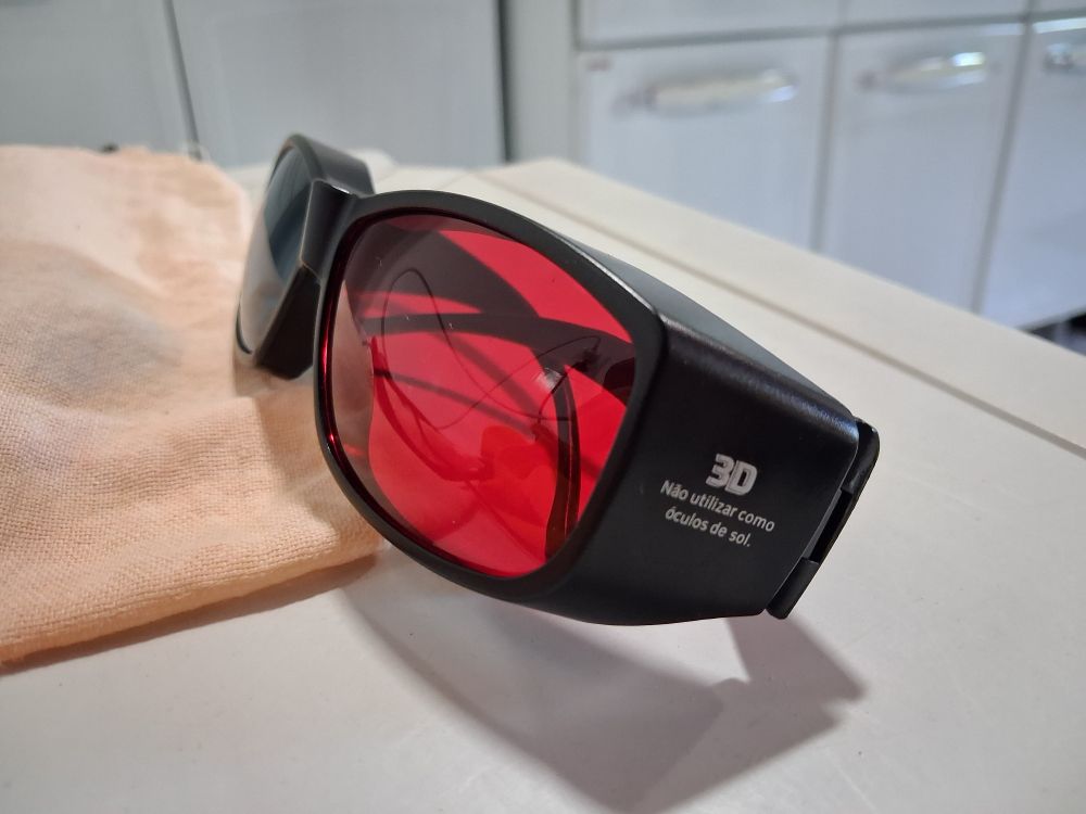 Vista lateral e frontal dos óculos 3D, com a lente vermelha em primeiro plano. O braço lateral direito mostra as inscrições "3D" e, abaixo, "Não utilizar como óculos de sol."