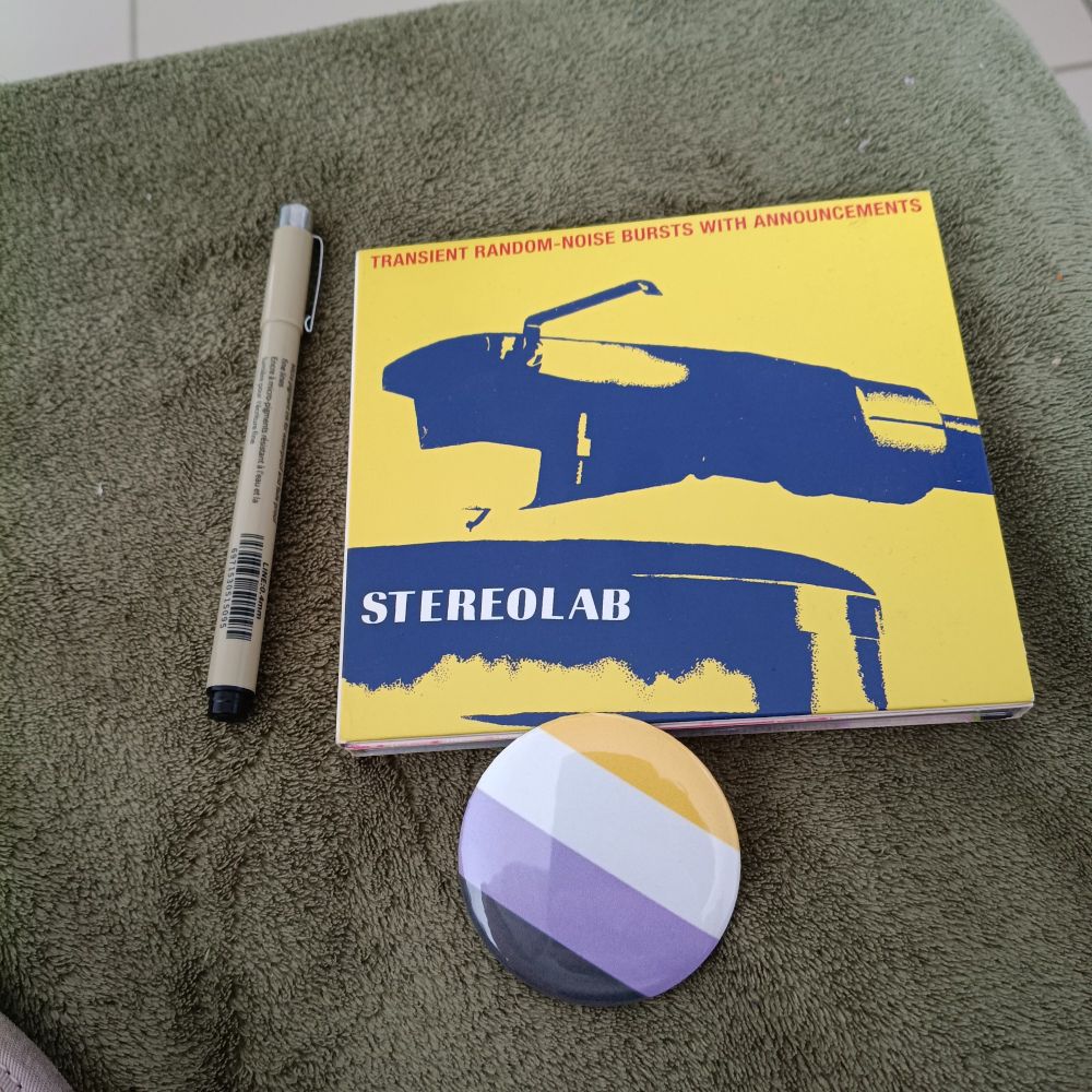 a pen, the enby flag pin and the expanded cd edition of stereolab's masterpiece, transient random-noise bursts with announcements.