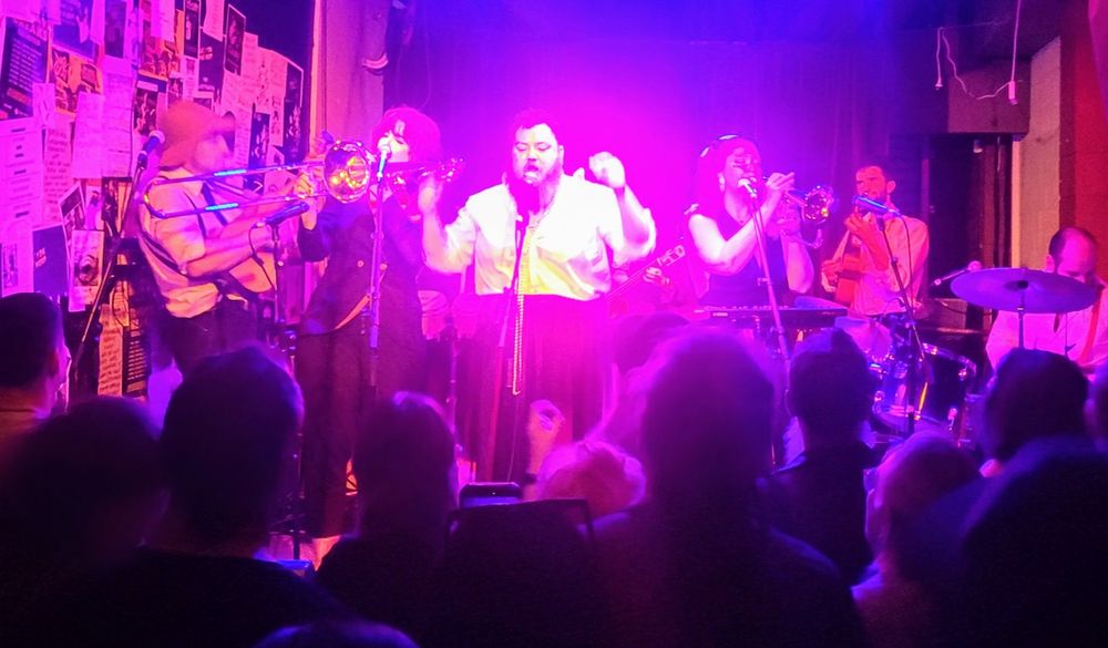A band badly photographed in red light with a lead singer two horn players and a ukulele 