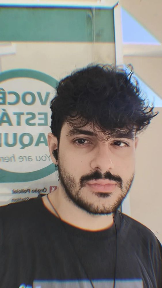 Selfie minha. Sou um homem branco de cabelos ondulados escuros e barba preta. Estou sem óculos e vestindo uma camisa preta com um colar no pescoço em frente a uma parada do BRT.