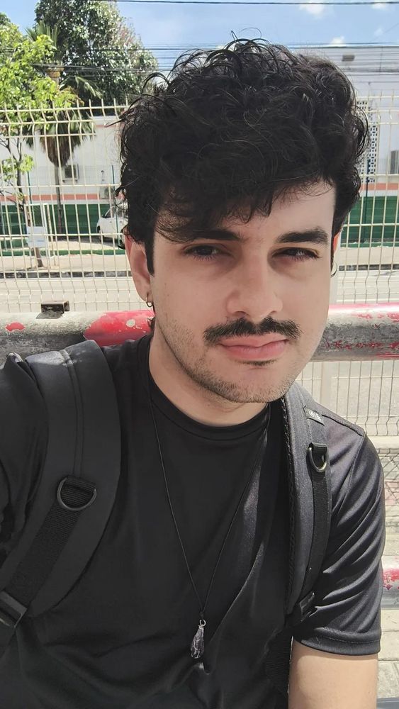 Selfie minha. Sou um homem branco de cabelos escuros cacheados, vestindo uma camisa preta com mochila e um colar de ametista. Tenho um bigode e estou sem óculos 