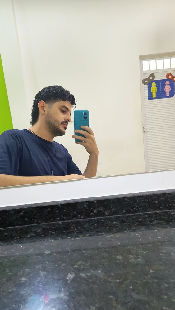 Selfie minha no espelho. Sou um homem branco de cabelo escuro em corte mullet, sem óculos e com uma barba rala. Visto uma camisa azul e bato foto de mim mesmo num espelho de lanchonete 