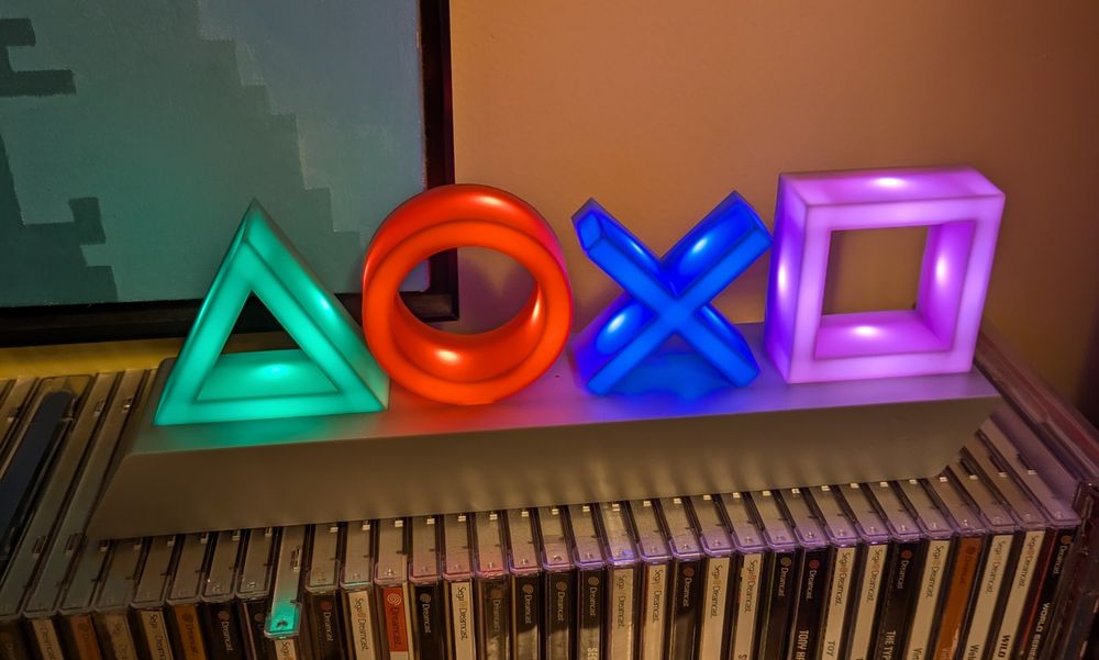 An illuminated PlayStation sign, it has a green triangle, a red circle, a blue X, and purple square, all lit by internal LED lights. It is sitting on top of a shelf of Sega Dreamcast games in jewel cases.