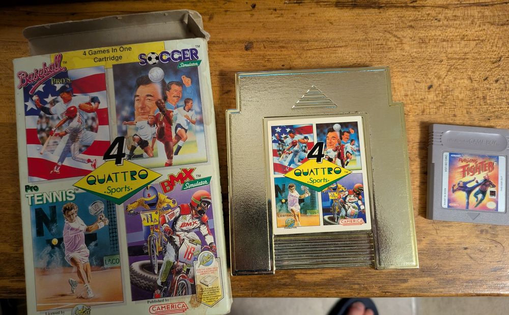 A complete-in-box copy of Quattro Sports for the NES. The box is laying next to the loose cartridge, which has a reflective gold finish. Next to them is a loose cartridge of Raging Fighter for the original Game Boy. The games are sitting on a wooden desk 