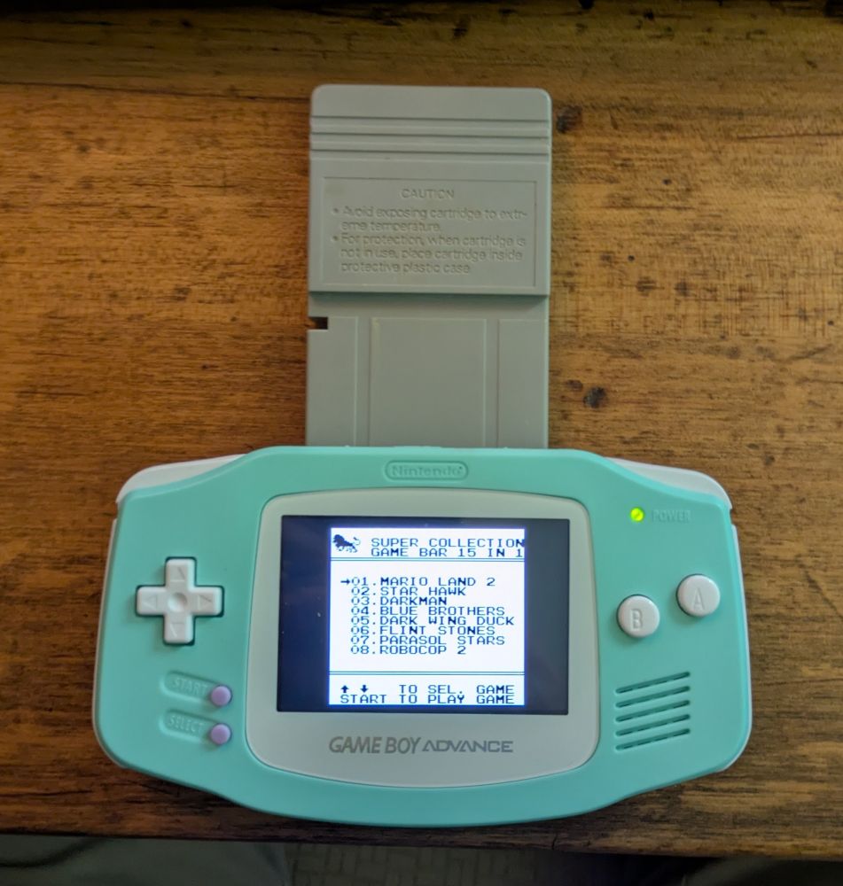 A double height bootleg Game Boy cartridge inserted into a Game Boy Advance. The extra height of the cartridge looks especially strange in the GBA. The screen shows the first eight games on the cartridge. In order, they are 1) Super Mario Land 2 2)Star Hawk 3) Darkman 4) Blue Brothers 5) Dark Wing Duck 6) Flint Stones 7) Parasol Stars and 8) Robocop 2.