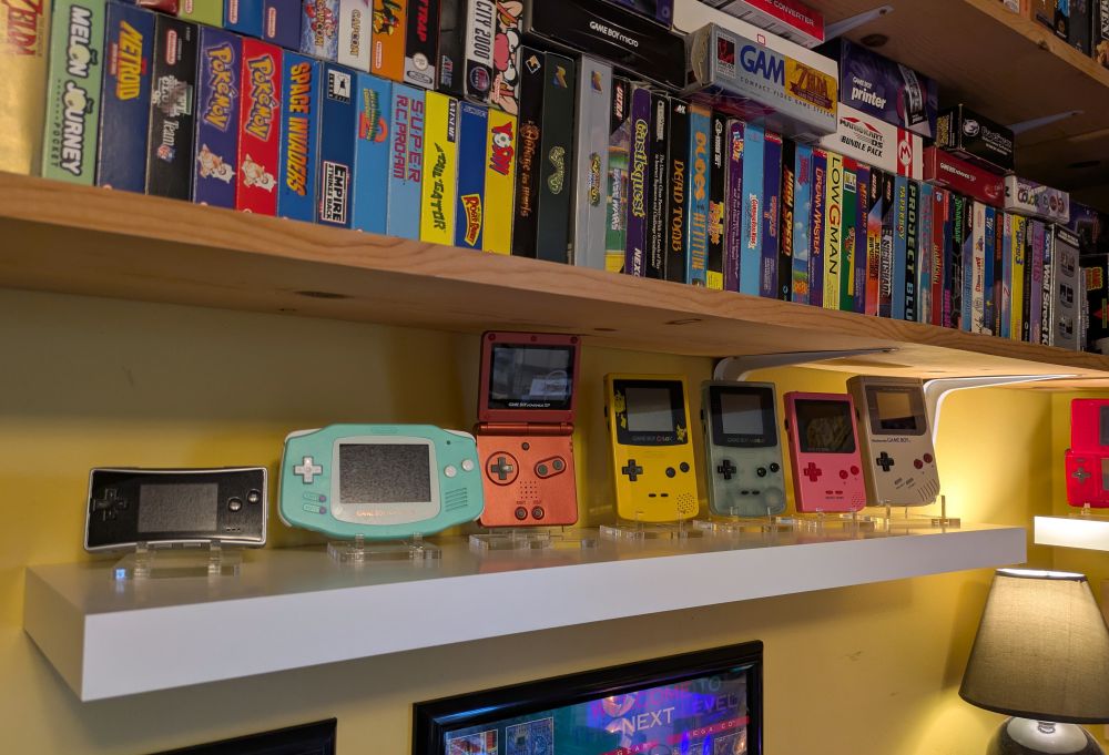 Shelving holding video game boxes, there's a white floating shelf at the bottom that has handheld consoles. From left to right is a Game Boy Micro, a Game Boy Advance with an aftermarket teal case and white buttons and lens, a flame red Game Boy Advance SP, a Game Boy Color in yellow with a Pikachu lens installed, a Game Boy Color with an aftermarket glow in the dark Game Boy Horror case, a pink Game Boy Pocket, and an original Game Boy.