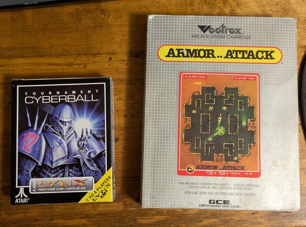 Two complete-in-box video games on a wooden table. The games are Tournament Cyberball for Atari Lynx and Armor..Attack for Vectrex.