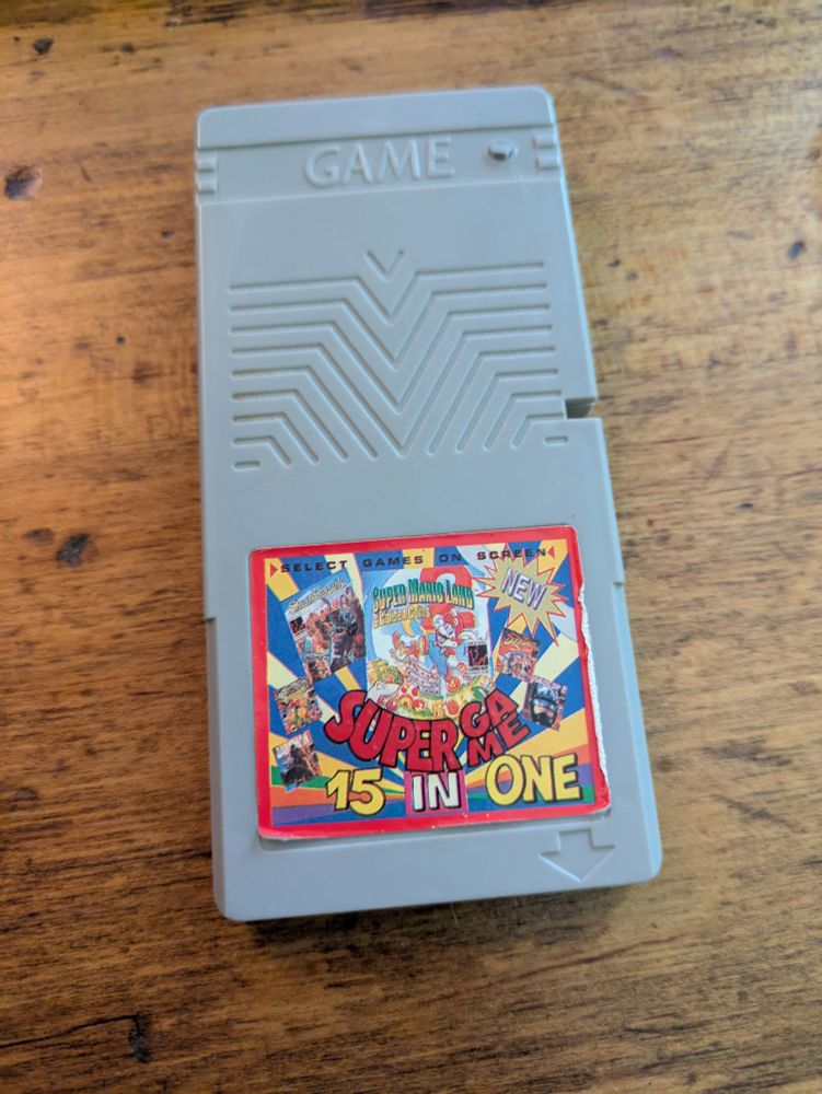 Bootleg Game Boy cartridge sitting on a wooden desk. The cart is twice as tall as a standard Game Boy cartridge. The label says SUPER GAME 15 IN 1 on it.