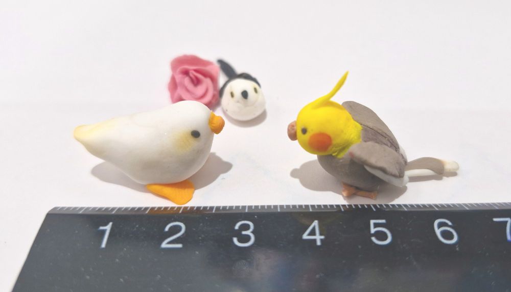 three polymer clay bird figurines next to a ruler. on the left is the budgie-duck hybrid with a cute yellow blush, approximately 2cm long. on the right is a cockatiel with heart wings approximately 2.5cm long. in the middle is a munted looking long tailed tit. next to it is a small rose. both of them are approximately 0.75cm long. 