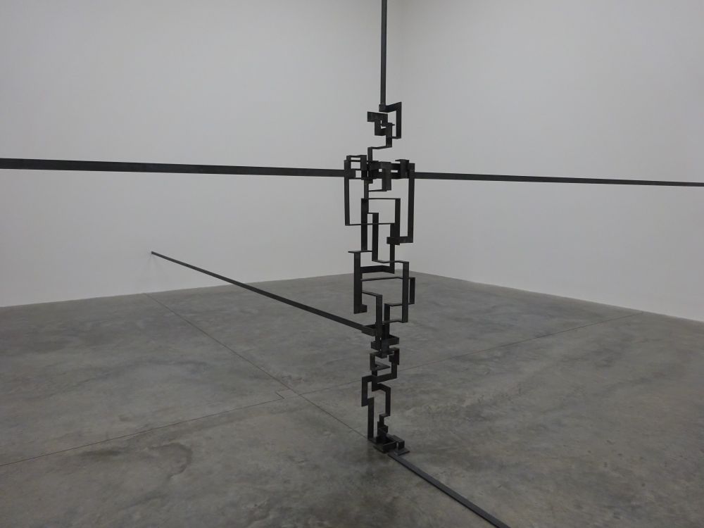 A sculpture made from five metal lines that come in from the walls and ceiling, and which are shaped in the middle to create the impression of a person standing.