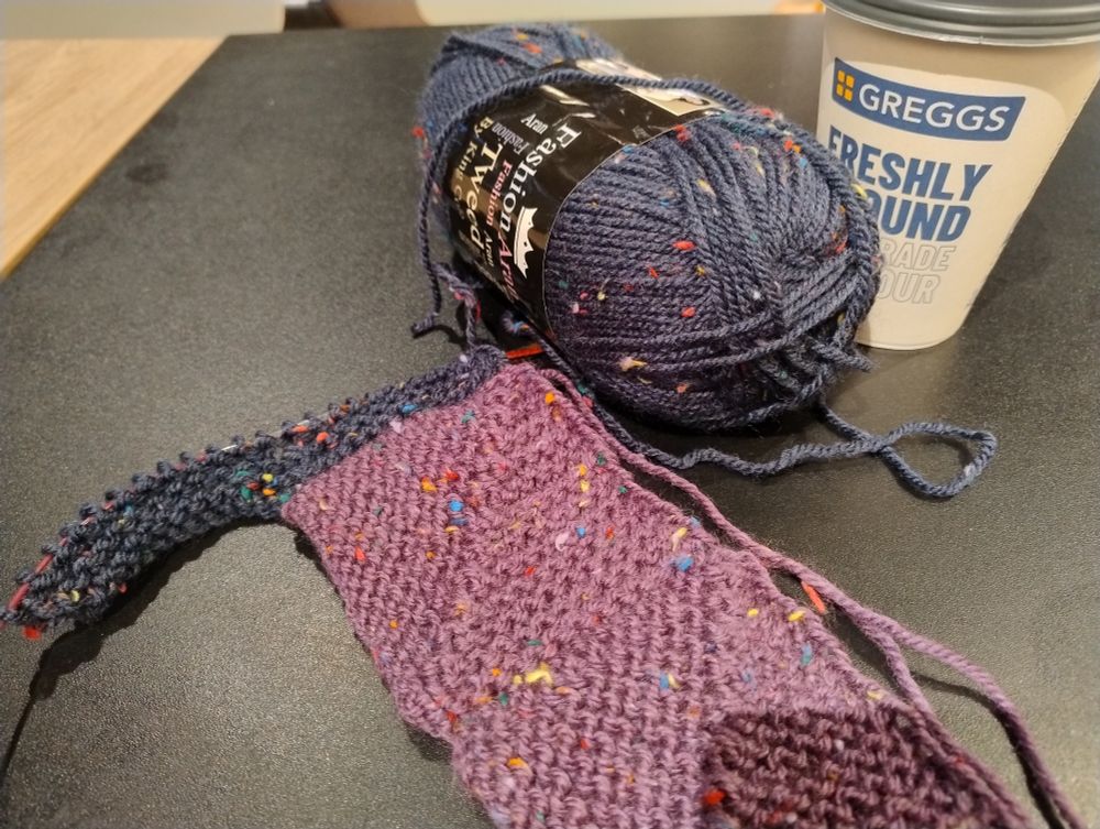 Mitered square knitting resting on a table next to a Greggs coffee.