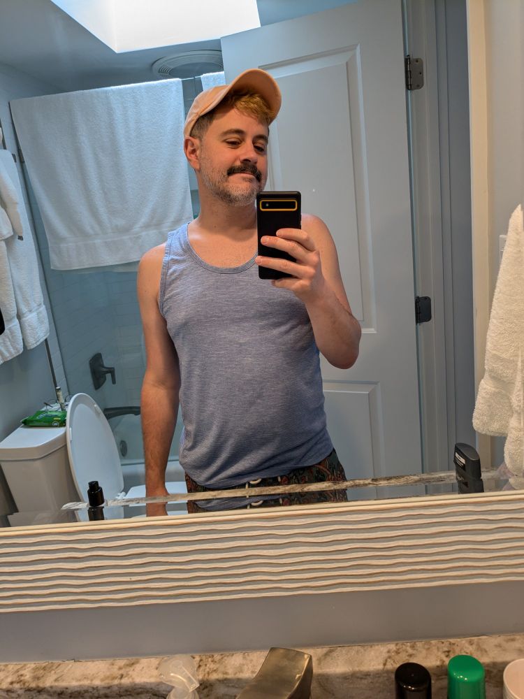 Mustachioed man takes a mirror selfie, he's wearing a peach-colored ballcap and a light blue tank top and his belly is bulging because he was on vacation and very full of food at the time and not because it's always like that :)