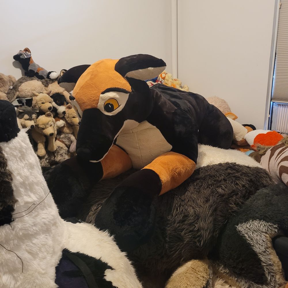 A giant PlushLife german shepherd plush lies atop an even bigger custom plush's tail in a room full of countless other canine plushies.