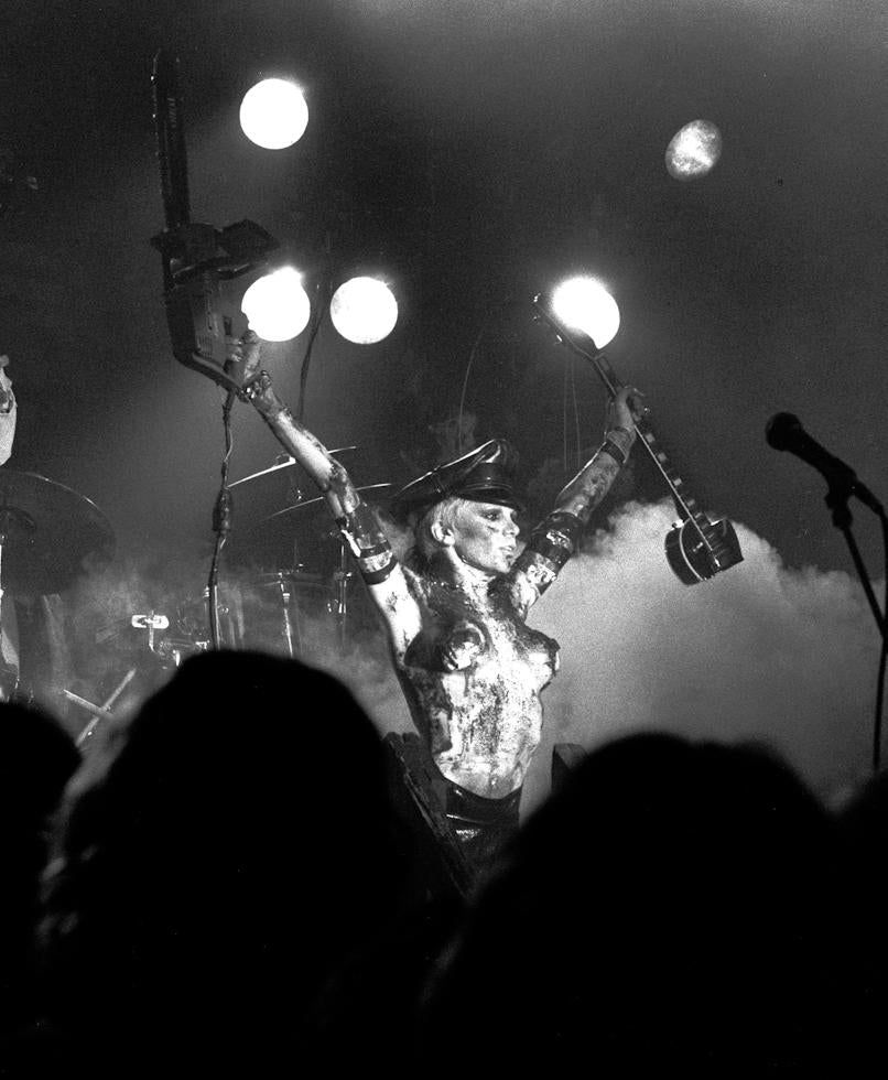 Black and white photo of The Queen of Shock Rock on stage with a chainsaw held aloft in her right hand and the remains of an electric guitar in her left.