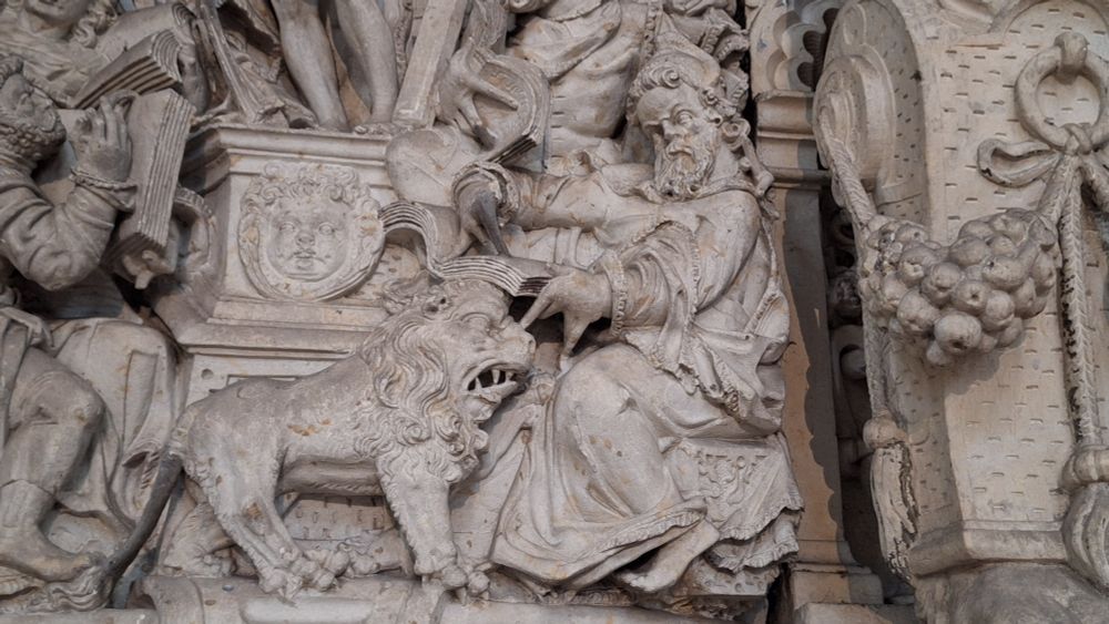 St. Jerome and the lion as part of a fresco in Magdeburg Cathedral.  St. Jerome is depicted sitting down and holding a book. He wears long garments and has a beard. His fingers are spread in unnatural ways. Next to him, there's a very stocky lion with its mouth open and its nose russled. St. Jerome's book rests on the lion's head. Both look immensly grumpy. 