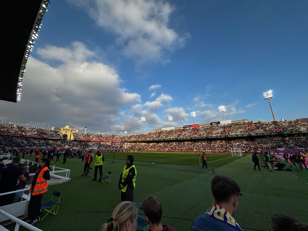 Photo taken from the corner of the Estadi Olímpic de Montjuïc as a field-side spectator before a soccer match. There are thousands of people in the stands and stadium security preventing onlookers from hopping the small fence onto the green soccer field. 