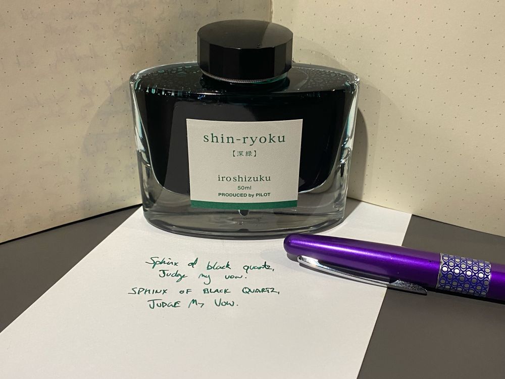 A pot of dark green ink sits on top of an envelope, and in front of a blank notebook. The label reads “shin-ryoku — iroshizuku” and describes a 50ml pot of ink. To the side sits a purple fountain pen with its lid closed. 

In green handwriting, both lowercase and uppercase letters spell “Sphinx of black quartz, judge my vow”. 