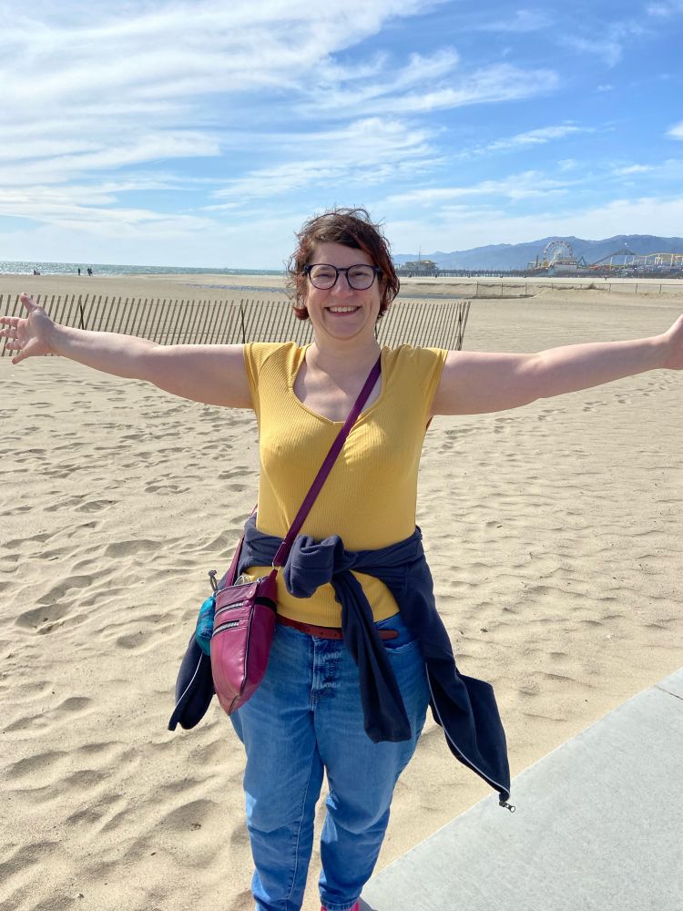 Sylvia, a middle-aged white lady with brown hair and glasses, stands where the path meets the sand at Santa Monica beach. She’s grinning and holding her arms straight out, like she wants to hug the beach. Behind her the same stretches towards the sea on the left and hills on the left.